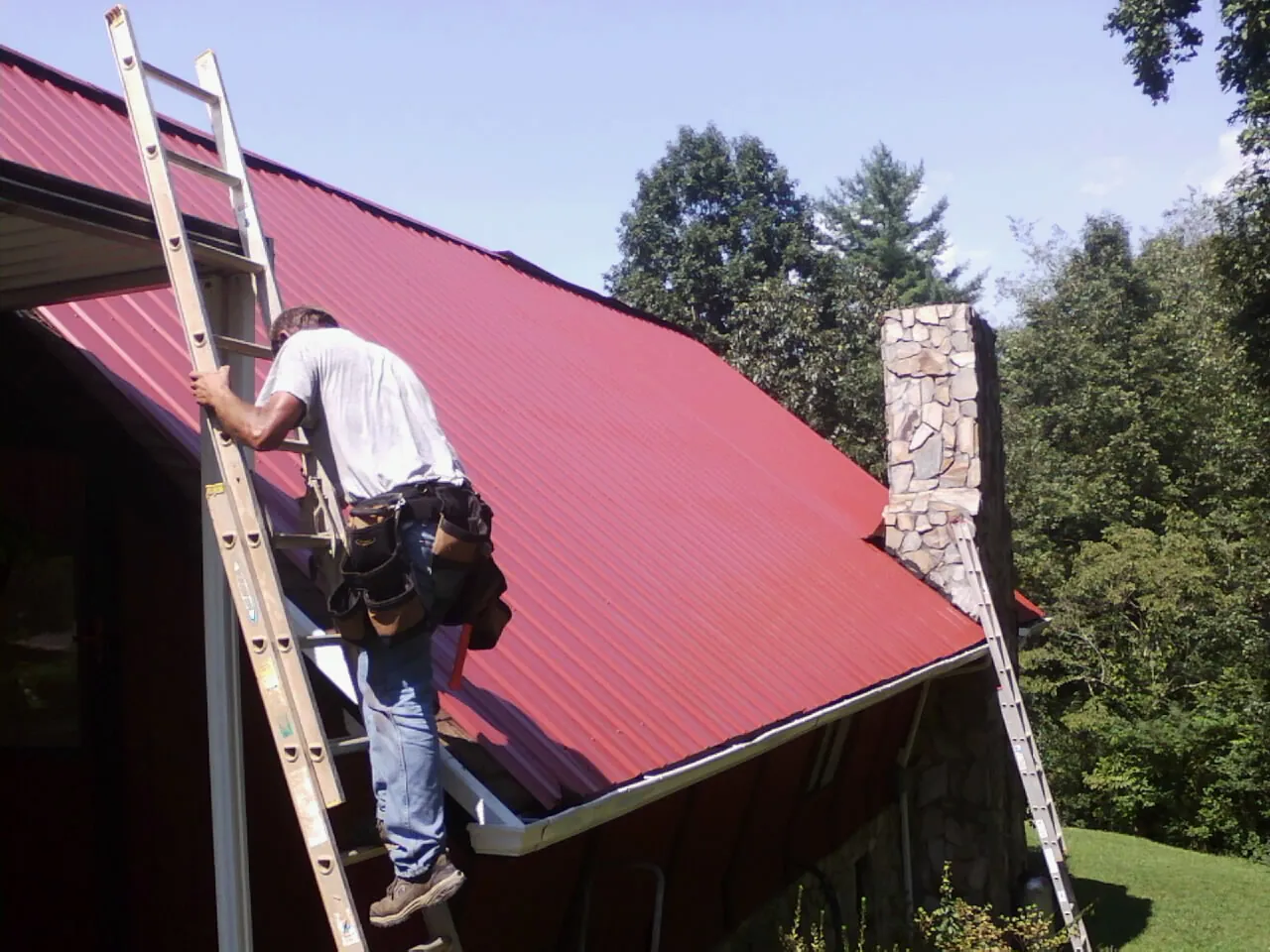 Roofer on a ladder beside a red metal roof and stone chimney — Soffit & Fascia Repair by Roofing Brothers in Baraboo